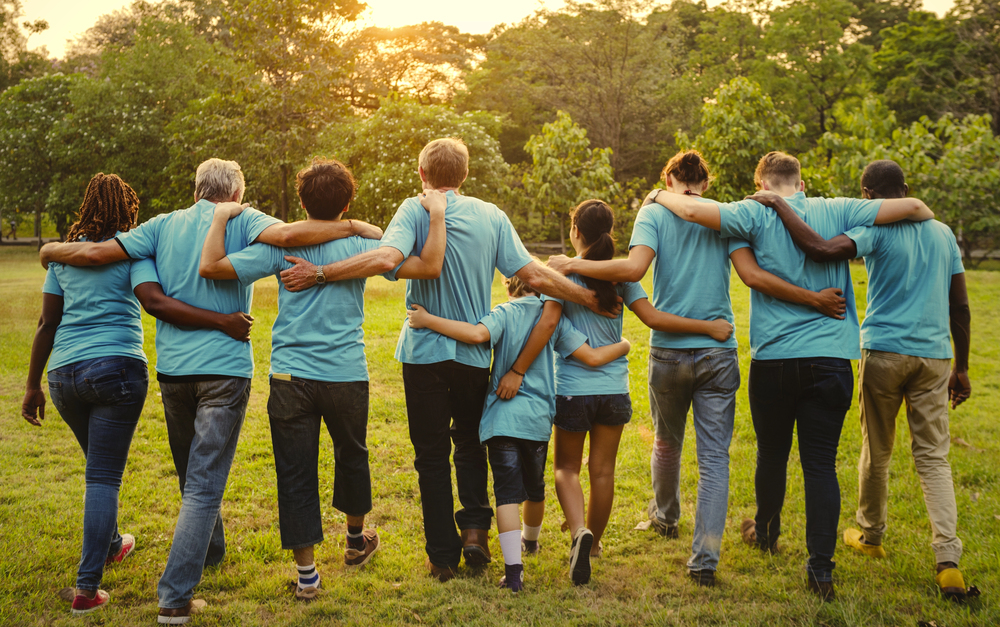 A group of people wearing blue t-shirts stand in a line with their arms wrapped around each others shoulders. The background features green trees and the people are standing on a green lawn.