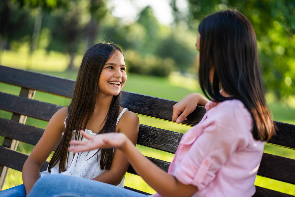 An adult woman chats with a young girl while sitting on a park bench.