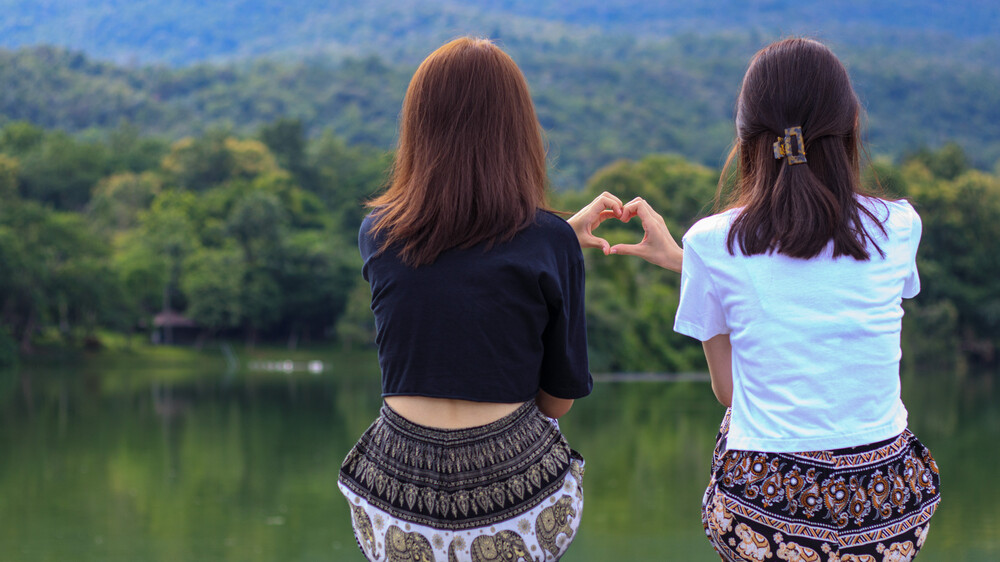 Two girls use their hands to create a heart symbol in front of beautiful green mountain landscape.